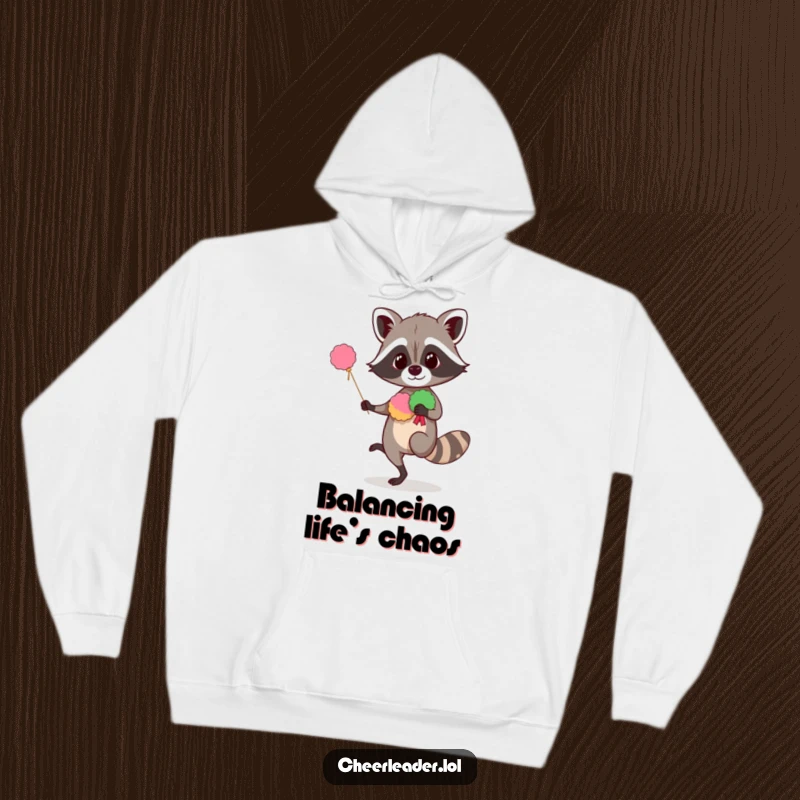 Funny raccoon hoodie showing a curious creature balancing on one leg with pom-poms, offering humorous warmth and a cheerful design.